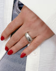 Hand wearing a silver Limestone Statement ring, featuring a smooth, rounded design. Red nail polish on almond-shaped nails contrasts with white clothing and gray fabric in the background, showcasing the ring's bold and elegant appearance.