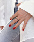 Hand wearing unique Sand Drip Band ring, showcasing delicate design inspired by ocean water drips. Contrasting with white shirt, blue jeans, and vibrant red nail polish, highlighting elegant jewelry piece.